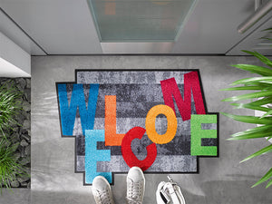 Bild in Slideshow öffnen, Fußmatte in spezieller Form mit bunten, tanzenden Buchstaben &quot;WELCOME&quot; vor der Eingangstür
